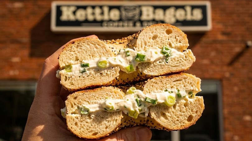 Hand holding a Kettle Bagels sesame bagel with scallion cream cheese, outside the shop.