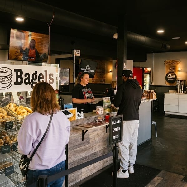 Customers ordering at the counter of Kettle Bagels, with a display of fresh bagels in the foreground.
