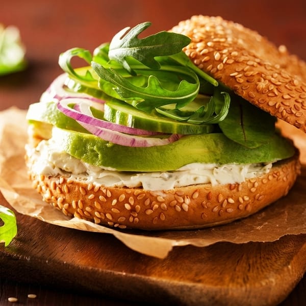 Sesame bagel sandwich with cream cheese, avocado, cucumber, red onion, and arugula on a wooden board.