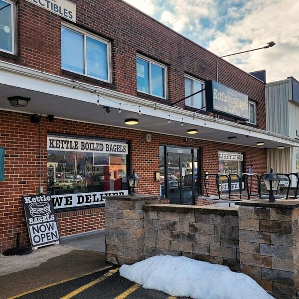 Exterior of Kettle Boiled Bagels, a brick building with a "Now Open" sign and snow on the ground.