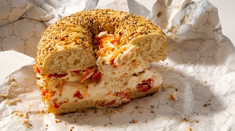 Everything bagel with cream cheese and sun-dried tomatoes, resting on crumpled white paper.